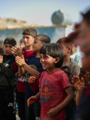 A group of children smiling and clapping during an outdoor celebration in Idlib, Syria.